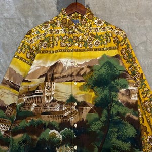 Puede incluir: Camisa amarilla con botones con un diseño de paisaje que presenta un castillo, montañas y árboles. El cuello y las mangas tienen un estampado floral en tonos amarillos, marrones y verdes. La camisa está hecha de una tela ligera.