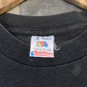 Może przedstawiać: Zbliżenie kołnierza czarnej koszulki z metką Fruit of the Loom. Etykieta jest biała z czerwonymi, niebieskimi i ż&oacute;łtymi grafikami i tekstem. Na etykiecie widnieje napis "100% Bawełna, kurczona" i "Made in USA".