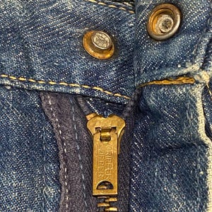 Może przedstawiać: Niebieska odzież dżinsowa z mosiężnym metalowym suwakiem z wytłoczonym napisem GRIPPER ZIPPER. Zapięcie zawiera metalowy guzik i zatrzask, oba z widocznymi śladami użytkowania, umieszczone na tle teksturowanego niebieskiego materiału.