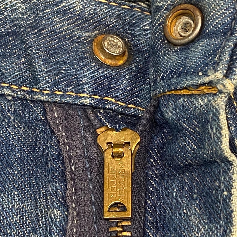 Może przedstawiać: Niebieska odzież dżinsowa z mosiężnym metalowym suwakiem z wytłoczonym napisem GRIPPER ZIPPER. Zapięcie zawiera metalowy guzik i zatrzask, oba z widocznymi śladami użytkowania, umieszczone na tle teksturowanego niebieskiego materiału.