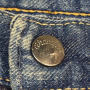 Może przedstawiać: Metalowy guzik z wytłoczonym napisem Wrangler przymocowany do niebieskiej tkaniny dżinsowej. Dżins ma wyraźny splot skośny i jest przeszyty grubą, kontrastującą ż&oacute;łtą nicią wzdłuż szw&oacute;w oraz wok&oacute;ł otworu na guzik.