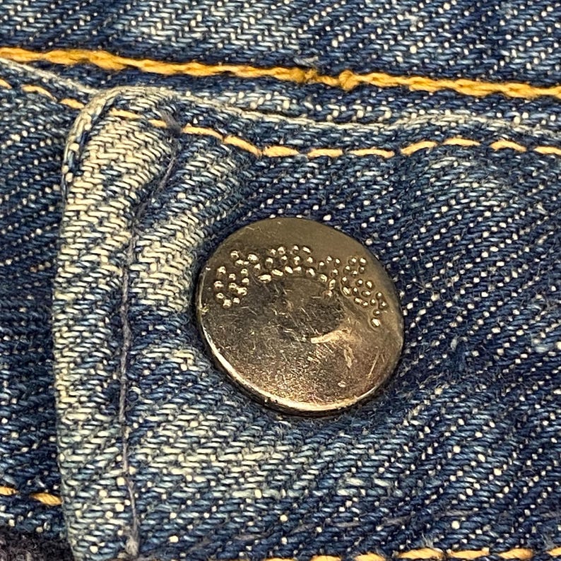 Może przedstawiać: Metalowy guzik z wytłoczonym napisem Wrangler przymocowany do niebieskiej tkaniny dżinsowej. Dżins ma wyraźny splot skośny i jest przeszyty grubą, kontrastującą ż&oacute;łtą nicią wzdłuż szw&oacute;w oraz wok&oacute;ł otworu na guzik.