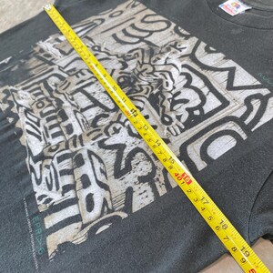 Może przedstawiać: Czarny t-shirt z biało-szarym wzorem graficznym. Ż&oacute;łta miarka wskazuje szerokość około 40 cm. Koszulka zawiera tekst "EPR6" i "1992 ANNIE LEIBOVITZ, FOTOFOLIO EDITIONS".