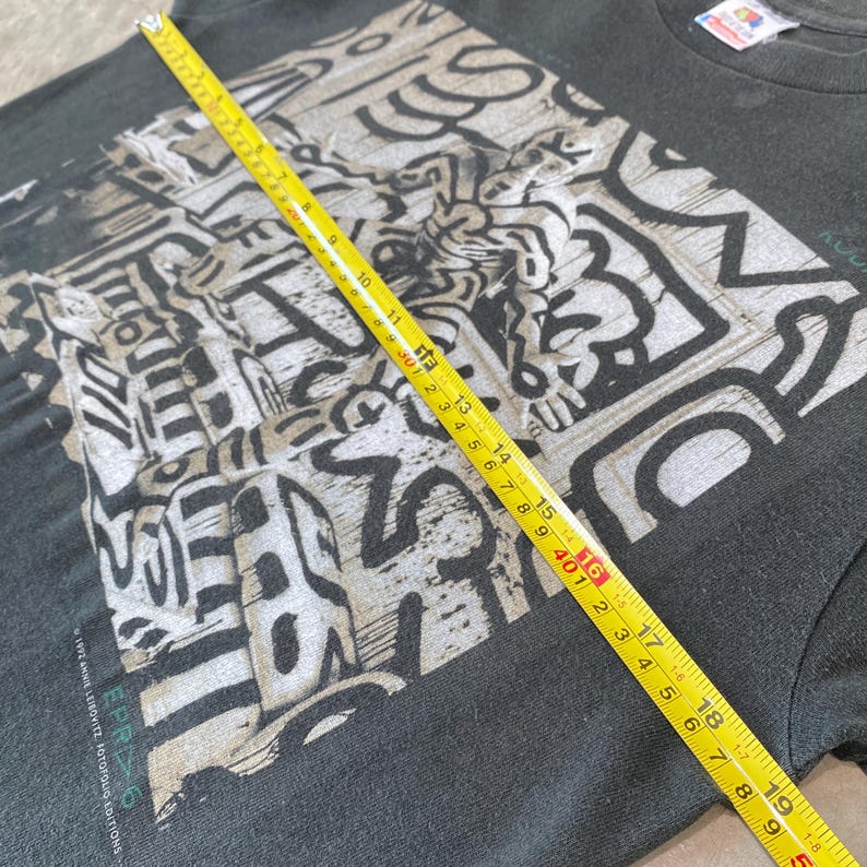 Może przedstawiać: Czarny t-shirt z biało-szarym wzorem graficznym. Ż&oacute;łta miarka wskazuje szerokość około 40 cm. Koszulka zawiera tekst "EPR6" i "1992 ANNIE LEIBOVITZ, FOTOFOLIO EDITIONS".