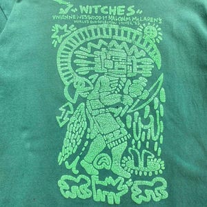 May include: Green t-shirt with a large, distressed green graphic. The graphic features a stylized figure with intricate details, surrounded by symbols and text that reads "Witches" and "Vivienne Westwood & Malcolm McLaren's World's End Collection LIVER '83."