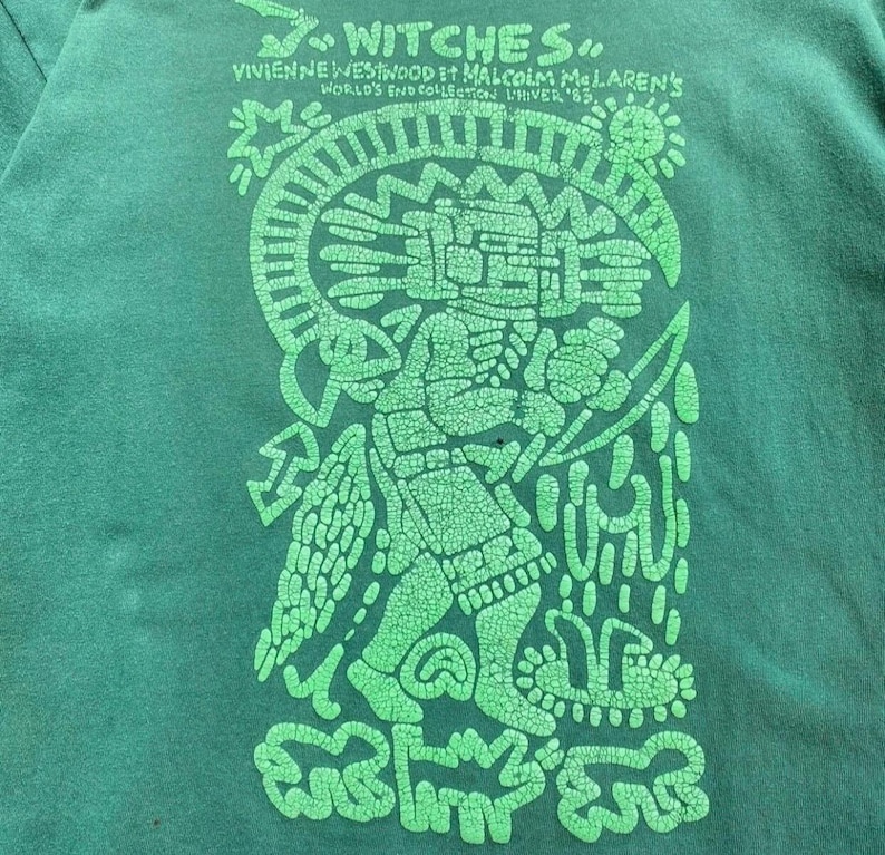 May include: Green t-shirt with a large, distressed green graphic. The graphic features a stylized figure with intricate details, surrounded by symbols and text that reads "Witches" and "Vivienne Westwood & Malcolm McLaren's World's End Collection LIVER '83."