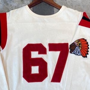 Könnte beinhalten: Cremefarbenes Sweatshirt im Vintage-Stil mit roten Akzenten und der Nummer 67. Auf dem Ärmel befindet sich ein Aufnäher mit einem indianischen Kopf. Die Ärmel haben rote Streifen. Das Sweatshirt hängt an einem Holzbügel.