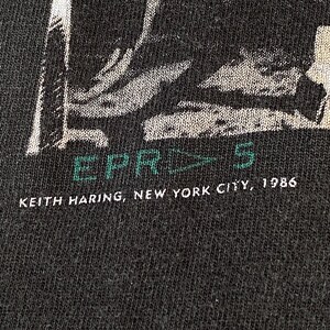 Może przedstawiać: Zbliżenie na ciemnoszarą koszulkę z graficznym wzorem. Tekst "EPR 5" jest w kolorze zielonym i białym. Poniżej tekst głosi "KEITH HARING, NEW YORK CITY, 1986" w kolorze białym.