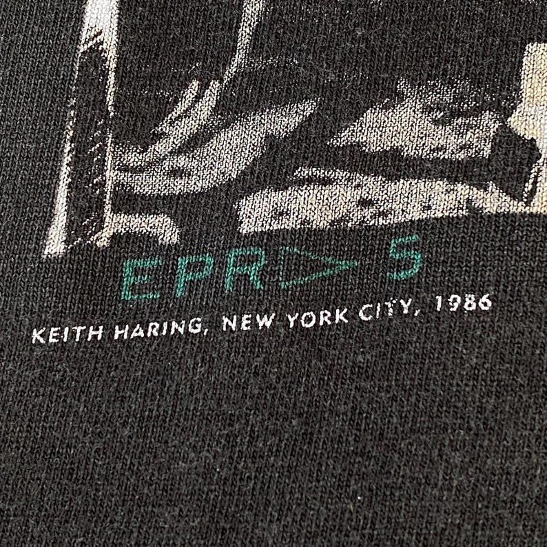 Może przedstawiać: Zbliżenie na ciemnoszarą koszulkę z graficznym wzorem. Tekst "EPR 5" jest w kolorze zielonym i białym. Poniżej tekst głosi "KEITH HARING, NEW YORK CITY, 1986" w kolorze białym.