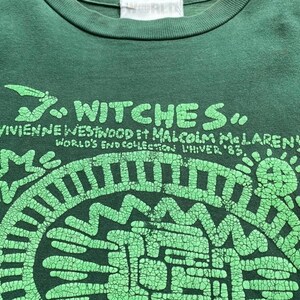 May include: A dark green t-shirt with a graphic design in light green. The design includes the text "WITCHES" above the text "VIVIENNE WESTWOOD et MALCOLM MCLAREN'S WORLD'S END COLLECTION UNIVER '83". The graphic is a circular pattern.