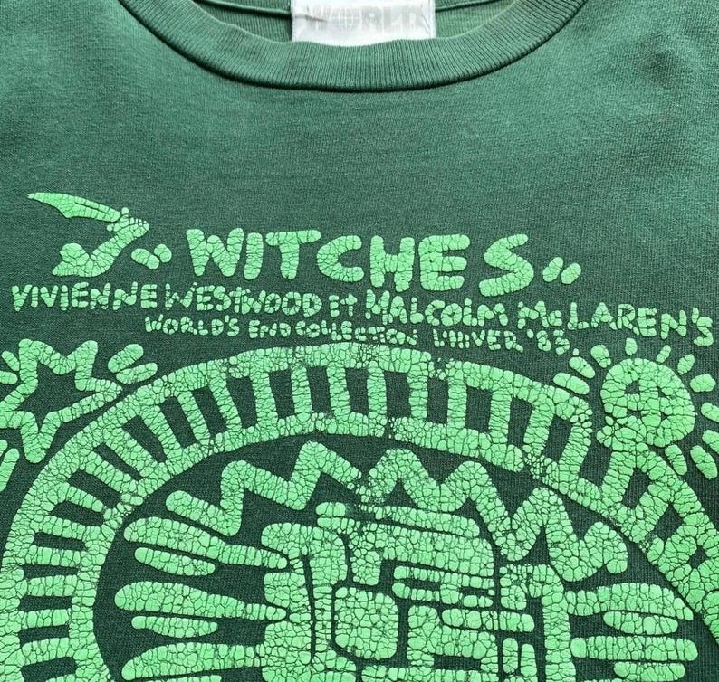 May include: A dark green t-shirt with a graphic design in light green. The design includes the text "WITCHES" above the text "VIVIENNE WESTWOOD et MALCOLM MCLAREN'S WORLD'S END COLLECTION UNIVER '83". The graphic is a circular pattern.