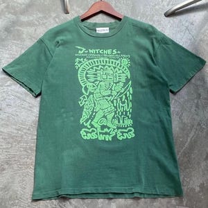 May include: Green t-shirt with a graphic design in neon green. The design features a stylized figure with text that reads "Witches" and other text. The shirt is hanging on a wooden hanger.