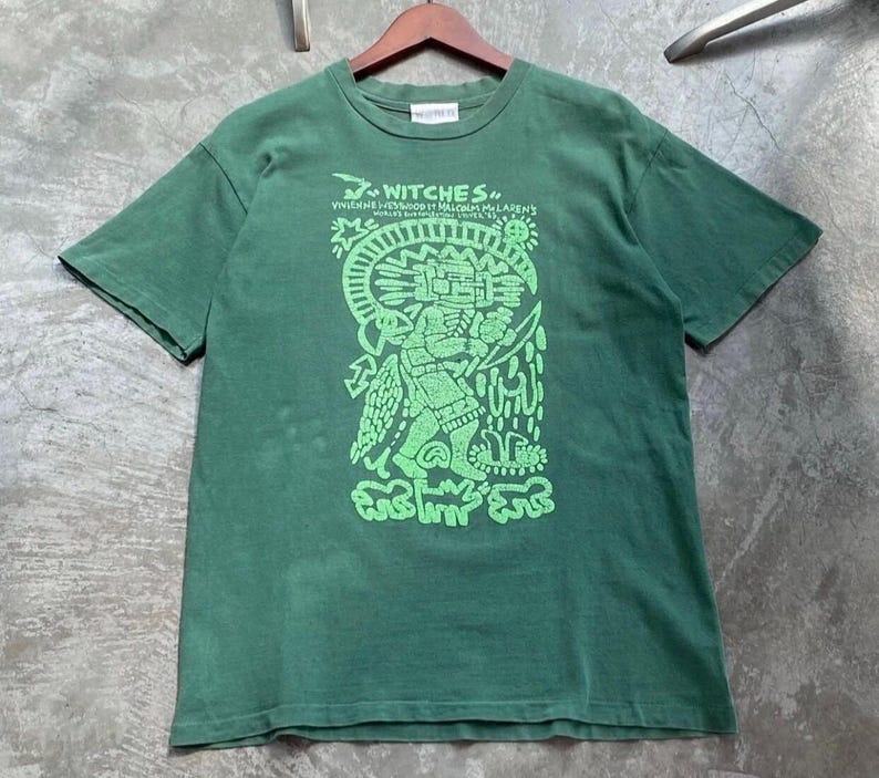 May include: Green t-shirt with a graphic design in neon green. The design features a stylized figure with text that reads "Witches" and other text. The shirt is hanging on a wooden hanger.