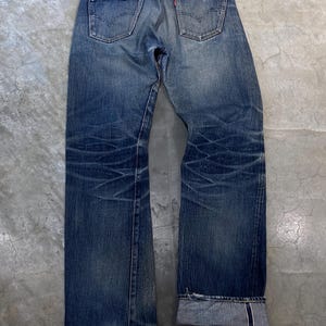 Puede incluir: Vaqueros de mezclilla azul descolorido con un diseño clásico de cinco bolsillos. Los vaqueros tienen un aspecto desgastado con decoloración y arrugas visibles. Los bolsillos traseros presentan un detalle de costura característico.