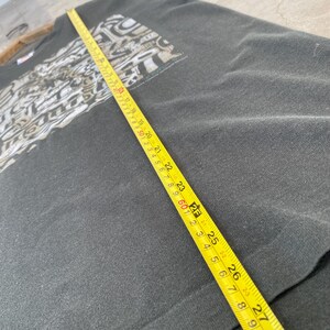 Może przedstawiać: Ciemnoszara koszulka z nadrukiem graficznym na g&oacute;rnej lewej części klatki piersiowej. Ż&oacute;łta miarka leży na koszulce, pokazując wymiary w centymetrach. Koszulka wydaje się być vintage.