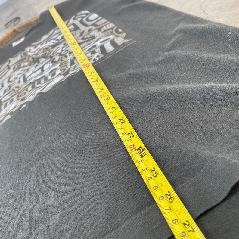 Może przedstawiać: Ciemnoszara koszulka z nadrukiem graficznym na g&oacute;rnej lewej części klatki piersiowej. Ż&oacute;łta miarka leży na koszulce, pokazując wymiary w centymetrach. Koszulka wydaje się być vintage.