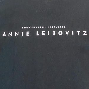 Może przedstawiać: Ciemnoszara okładka książki z białym napisem "ANNIE LEIBOVITZ". Nad imieniem znajduje się napis "PHOTOGRAPHS 1970-1990", r&oacute;wnież w kolorze białym. Tło jest jednolite, teksturowane i ciemnoszare.