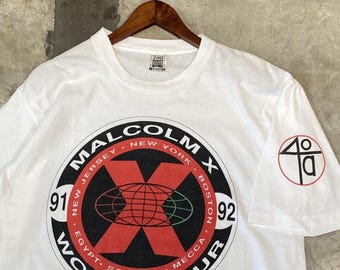 Pode incluir: Camiseta branca com um grande gráfico circular com o texto "MALCOLM X WORLD TOUR". O gráfico inclui um "X" vermelho sobre um globo, com nomes de cidades como Nova York e Boston. A manga tem um pequeno logotipo vermelho e preto.