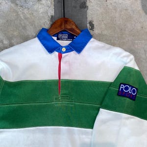 May include: A long-sleeved Polo Sport shirt with a white body, green horizontal stripe, and blue collar. The shirt features a red placket and the Polo Sport logo on the sleeve. The collar has the Polo by Ralph Lauren logo.