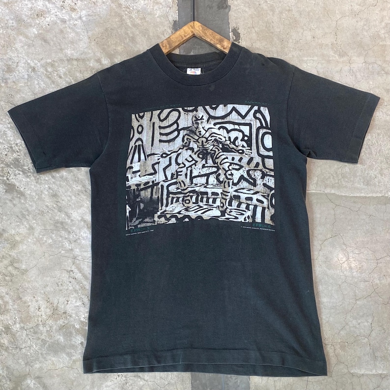 Może przedstawiać: Czarny t-shirt z kr&oacute;tkim rękawem z nadrukiem graficznym. Nadruk przedstawia postać z abstrakcyjnymi kształtami w kolorze białym i szarym. Koszulka wisi na drewnianym wieszaku.