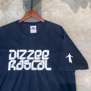 Può includere: T-shirt nero con la scritta bianca "Dizzee Rascal". Una silhouette bianca di una persona è sulla manica destra. La maglietta è appesa a un appendiabiti di legno.