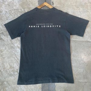 Może przedstawiać: Ciemnoszara koszulka z białym napisem "PHOTOGRAPHS 1970-1990 ANNIE LEIBOVITZ". Koszulka z kr&oacute;tkim rękawem ma okrągły dekolt i wisi na drewnianym wieszaku.