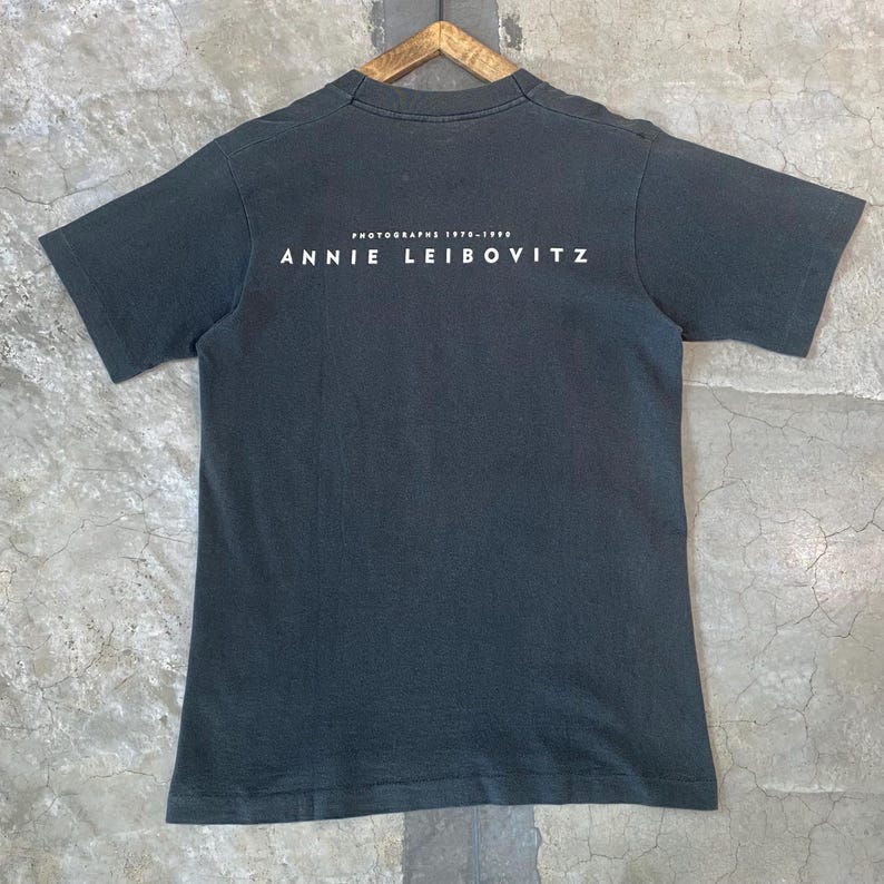 Może przedstawiać: Ciemnoszara koszulka z białym napisem "PHOTOGRAPHS 1970-1990 ANNIE LEIBOVITZ". Koszulka z kr&oacute;tkim rękawem ma okrągły dekolt i wisi na drewnianym wieszaku.