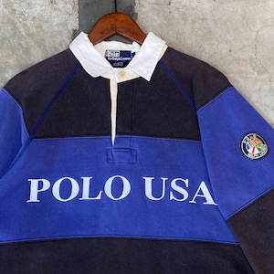 May include: A long-sleeve rugby shirt featuring a color-blocked design in black and blue. The shirt has a white collar, a button placket, and the text POLO USA printed in white across the chest. A circular crest patch is on the left sleeve.