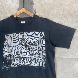 Może przedstawiać: Czarny t-shirt z kr&oacute;tkim rękawem z nadrukiem graficznym w kolorze białym i czarnym. Grafika przedstawia postać z odważnymi liniami i wzorami, z tekstem "KEITH HARING, NEW YORK CITY, 1986" na dole.