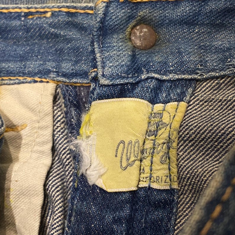 Może przedstawiać: Niebieskie dżinsy z metalowym guzikiem w talii oraz ż&oacute;łtą metką z tkaniny przyszytą przy rozporku. Na metce widnieje napis Wrangler oraz Sanforized wykonany czarną czcionką, a krawędź materiału jest wyraźnie postrzępiona i uszkodzona.
