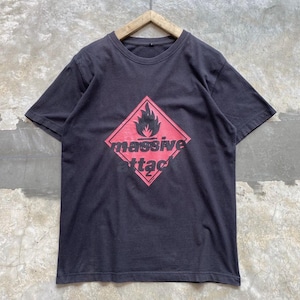 Puede incluir: Una camiseta negra de manga corta con un gráfico de rombo rojo en el pecho. Dentro del rombo hay un icono de llama negra sobre el texto "massive attack" impreso en letras minúsculas negras. Tiene cuello redondo y mangas cortas.