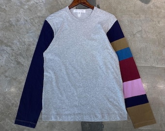 May include: A gray long-sleeve shirt with a navy blue sleeve on the left and a striped sleeve on the right. The stripes include navy, brown, blue, red, pink, and brown. The shirt is hanging on a wooden hanger.