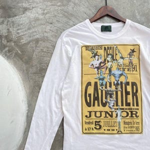 May include: White long-sleeve t-shirt with a vintage-style graphic. The graphic is a yellow poster with the word "GAUTIER" in large black letters, and the text "JUNIOR" below. The poster also includes the date "5 JUILLET 1991".