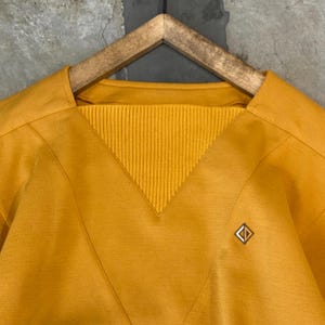 May include: Mustard yellow top with a V-neck and ribbed knit detail. A small, square gold-tone pin is on the right side. The top is displayed on a wooden hanger. The background is a neutral grey.