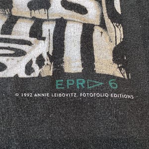 Może przedstawiać: Zbliżenie na ciemnoszarą koszulkę z wyblakłym wzorem graficznym. Tekst "EPR▷6" jest w kolorze turkusowym, a informacja o prawach autorskich brzmi "&copy; 1992 ANNIE LEIBOVITZ. FOTOFOLIO EDITIONS" w kolorze białym.