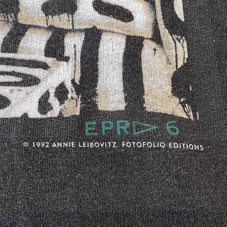Może przedstawiać: Zbliżenie na ciemnoszarą koszulkę z wyblakłym wzorem graficznym. Tekst "EPR▷6" jest w kolorze turkusowym, a informacja o prawach autorskich brzmi "&copy; 1992 ANNIE LEIBOVITZ. FOTOFOLIO EDITIONS" w kolorze białym.