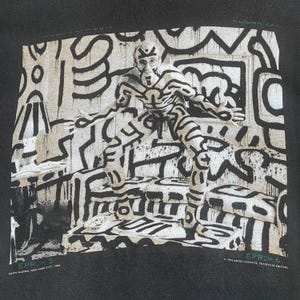 Może przedstawiać: Czarna bluza z nadrukiem Keitha Haringa. Grafika przedstawia osobę pokrytą białymi liniami i kształtami, stojącą w pokoju z podobną sztuką abstrakcyjną. Widoczny jest tekst "EPR>5" i "EPR>6", a także nazwisko artysty i rok 1986.