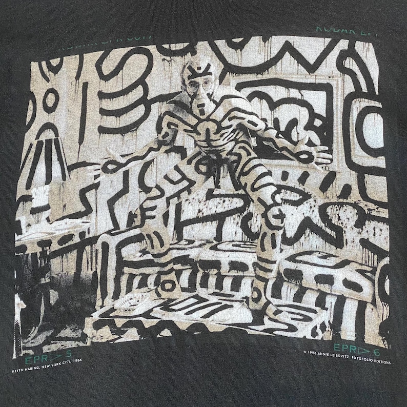 Może przedstawiać: Czarna bluza z nadrukiem Keitha Haringa. Grafika przedstawia osobę pokrytą białymi liniami i kształtami, stojącą w pokoju z podobną sztuką abstrakcyjną. Widoczny jest tekst "EPR>5" i "EPR>6", a także nazwisko artysty i rok 1986.