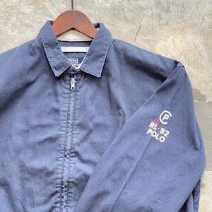 Puede incluir: Chaqueta azul marino con cuello y cremallera. El texto "RL-92 POLO" está en blanco en la manga izquierda. La chaqueta está colgada en una percha de madera.