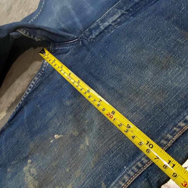 Może przedstawiać: Niebieskie spodnie dżinsowe z przetarciami i jasnymi odpryskami farby. Ż&oacute;łta taśma miernicza jest umieszczona ukośnie na materiale, wskazując długość około 29,8 cm wzdłuż mierzonego odcinka.