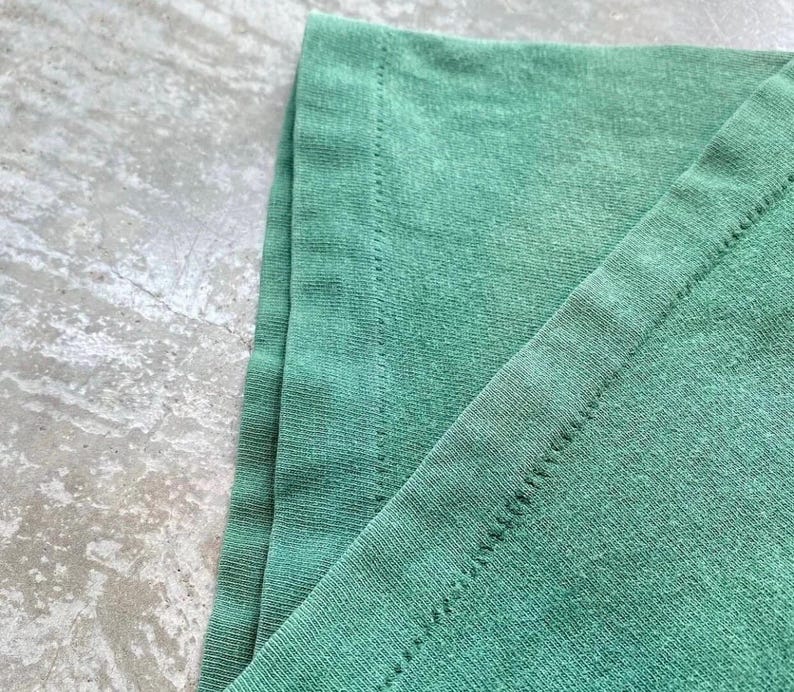 May include: Close-up of a section of a green t-shirt. The fabric is slightly faded, with visible stitching along the edges. The texture of the material is evident, showing the weave of the fibers.