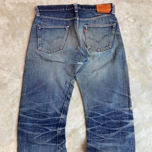 Op de afbeelding: Verbleekte blauwe denim jeans met een versleten uiterlijk. De jeans hebben een klassieke vijf-zakken-stijl, een rood logo-label en een distressed look met zichtbare slijtagepatronen. De achterzakken hebben een kenmerkend stikselontwerp.