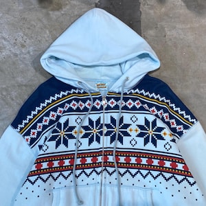 Puede incluir: Una sudadera con capucha azul claro con cremallera y un diseño estampado. El diseño presenta un canesú azul marino con un patrón geométrico en rojo, amarillo y blanco. La sudadera tiene una capucha con cordón y una pequeña etiqueta cerca de la capucha.