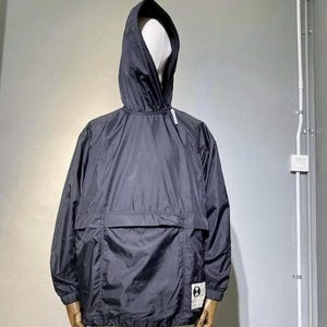Peut inclure: Anorak à capuche bleu marine foncé avec une grande poche avant. La veste a une capuche à cordon et une petite étiquette blanche sur le côté inférieur droit. L'anorak est présenté sur un mannequin.