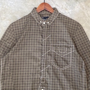 May include: Brown button-down shirt with a geometric pattern. The shirt features a single chest pocket and white stitching details. The shirt is hanging on a wooden hanger.