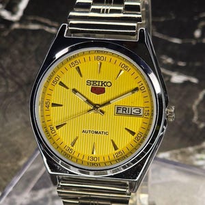 Op de afbeelding: Een zilverkleurig Seiko automatisch polshorloge met een heldergele wijzerplaat. De wijzerplaat heeft zwarte uurmarkeringen, een dag-datumweergave en het Seiko-logo. De horlogeband is gemaakt van zilverkleurige metalen schakels.
