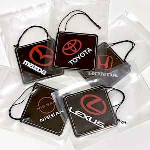 May include: A selection of car air fresheners with logos for Mazda, Toyota, Honda, Nissan, and Lexus. Each square freshener is black with a red or white logo, and a black string. They are packaged in clear plastic.
