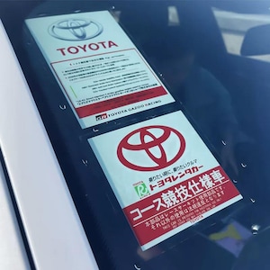 May include: Two white and red Toyota stickers are visible through a car windscreen. The top sticker has the Toyota logo and text. The bottom sticker has the Toyota logo and Japanese text, including "Toyota Rent-a-car".