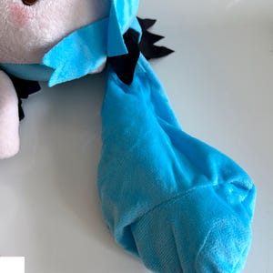 May include: A plush doll with a light blue and white color scheme. The doll has a blue hair accessory and black accents. The doll's face has brown eyes and pink cheeks. The doll is made of soft, plush material.