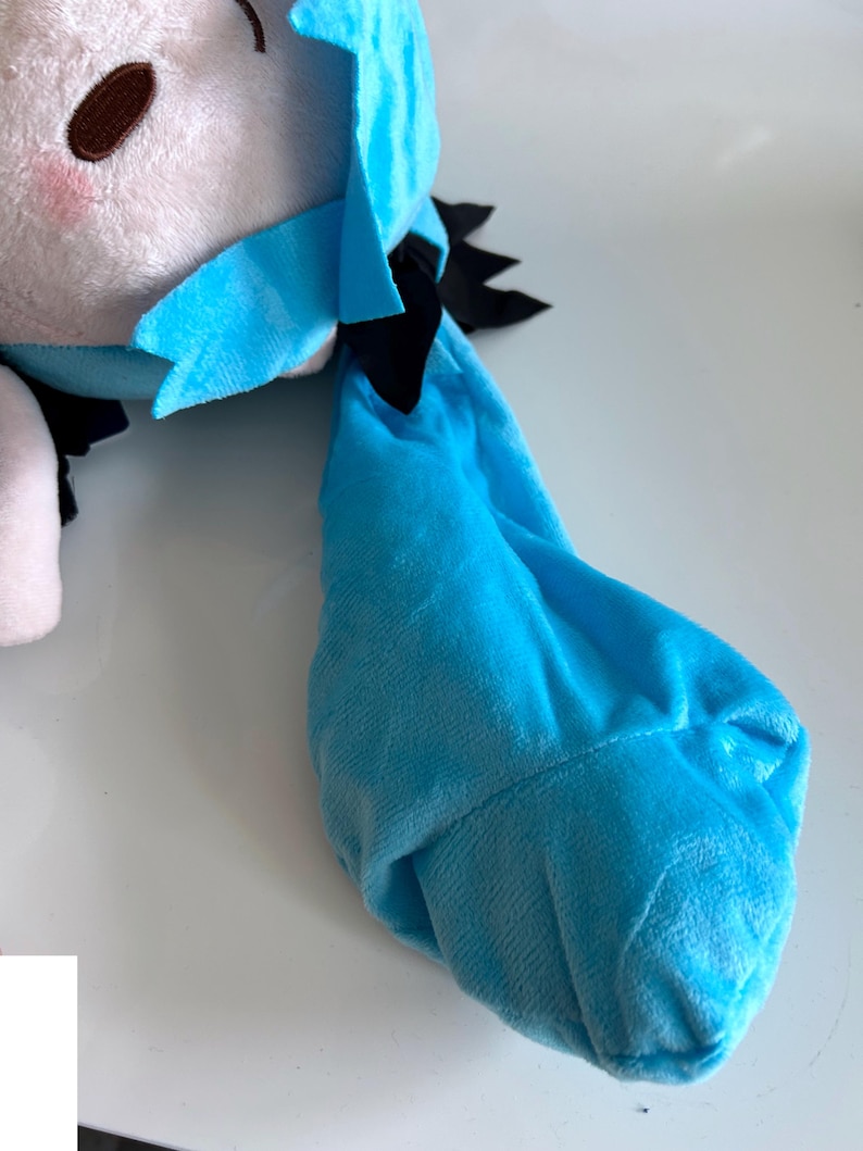 May include: A plush doll with a light blue and white color scheme. The doll has a blue hair accessory and black accents. The doll's face has brown eyes and pink cheeks. The doll is made of soft, plush material.